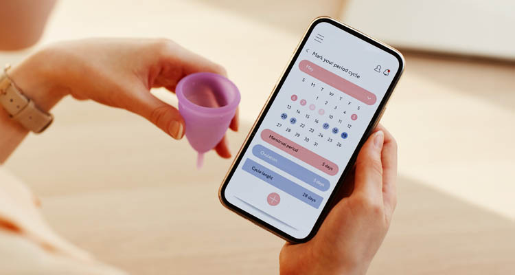 Eine Person hält eine violette Menstruationstasse in der Hand und schaut auf ein Smartphone mit geöffneter Perioden-Tracking-App.